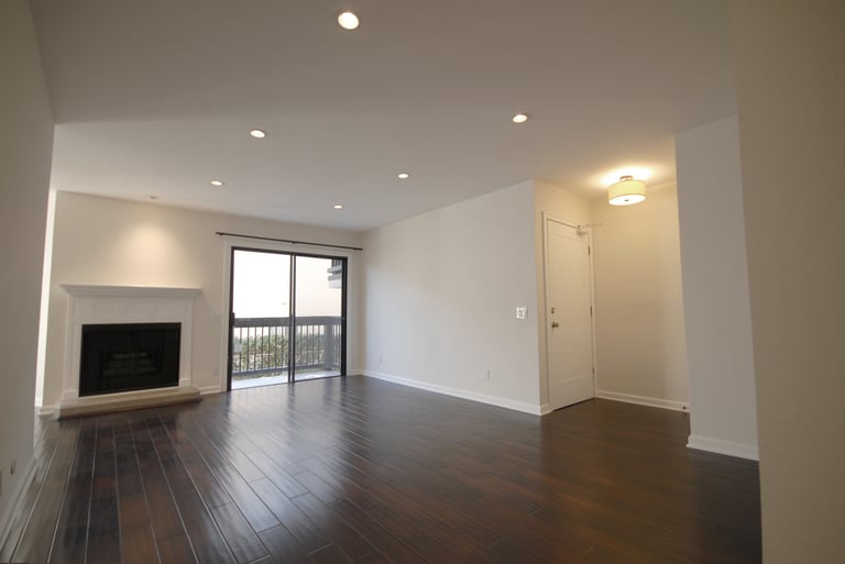 Modern empty living room with dark hardwood floors, recessed ceiling lights, mounted TV, and glass balcony doors