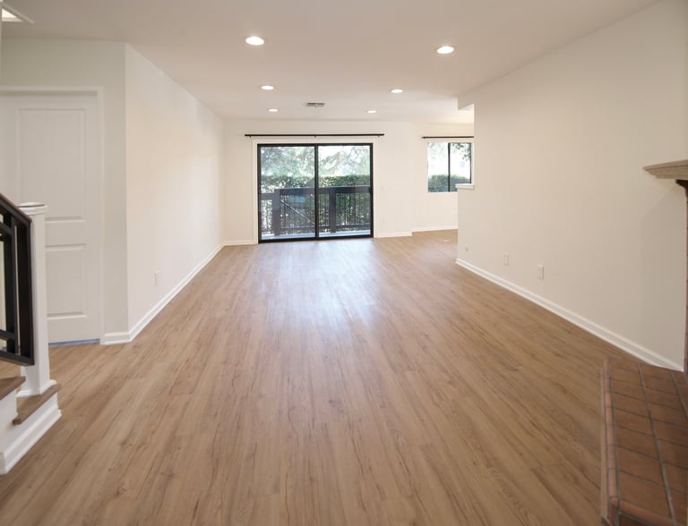 Empty modern living room with wood flooring, white walls, recessed lighting, and glass patio doors overlooking a balcony