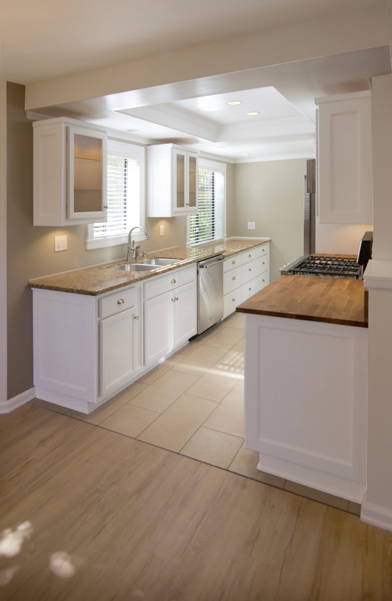 Modern galley kitchen with white cabinets, granite countertops, stainless steel sink, and island, illuminated by recessed ceiling lights