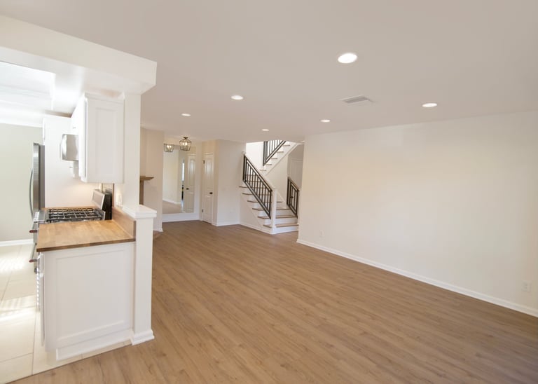 Modern kitchen and open living room with white cabinetry, stainless steel appliances, wood flooring, and staircase